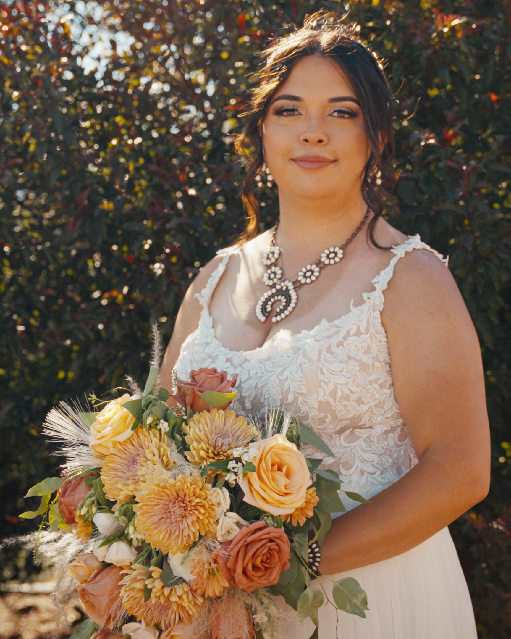 Bride in a lace wedding dress holding a bouquet of yellow and peach flowers outdoors.