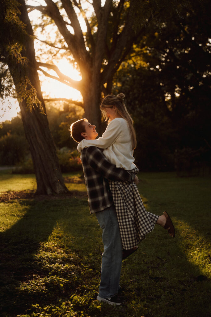 Couple laughing as one partner lifts the other beneath a sunlit tree.