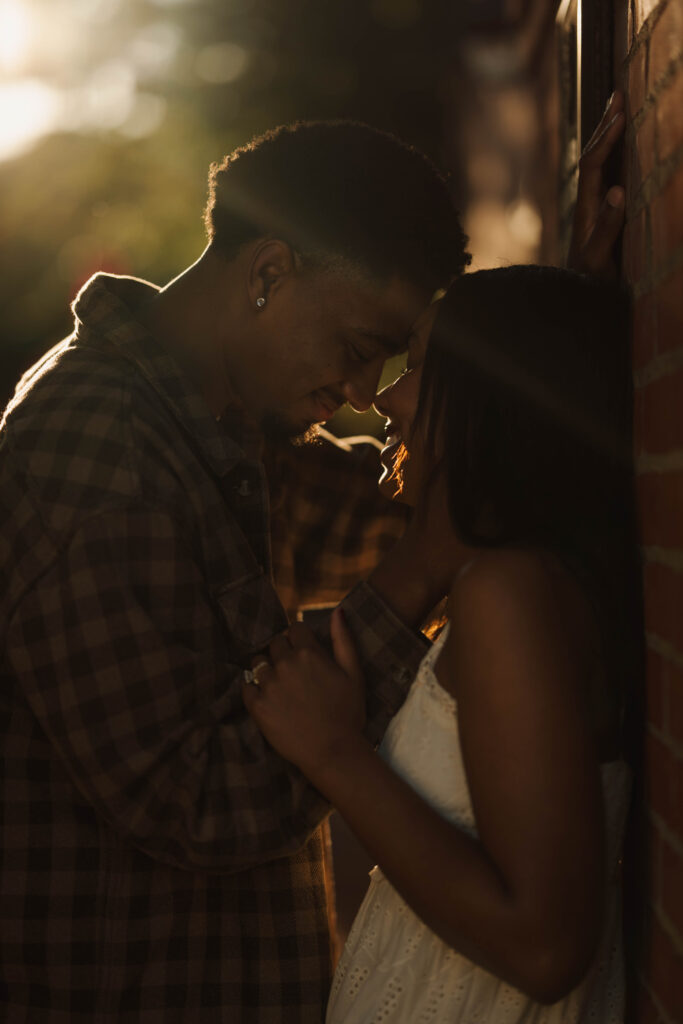 Couple touching foreheads beside a brick wall in warm backlight.