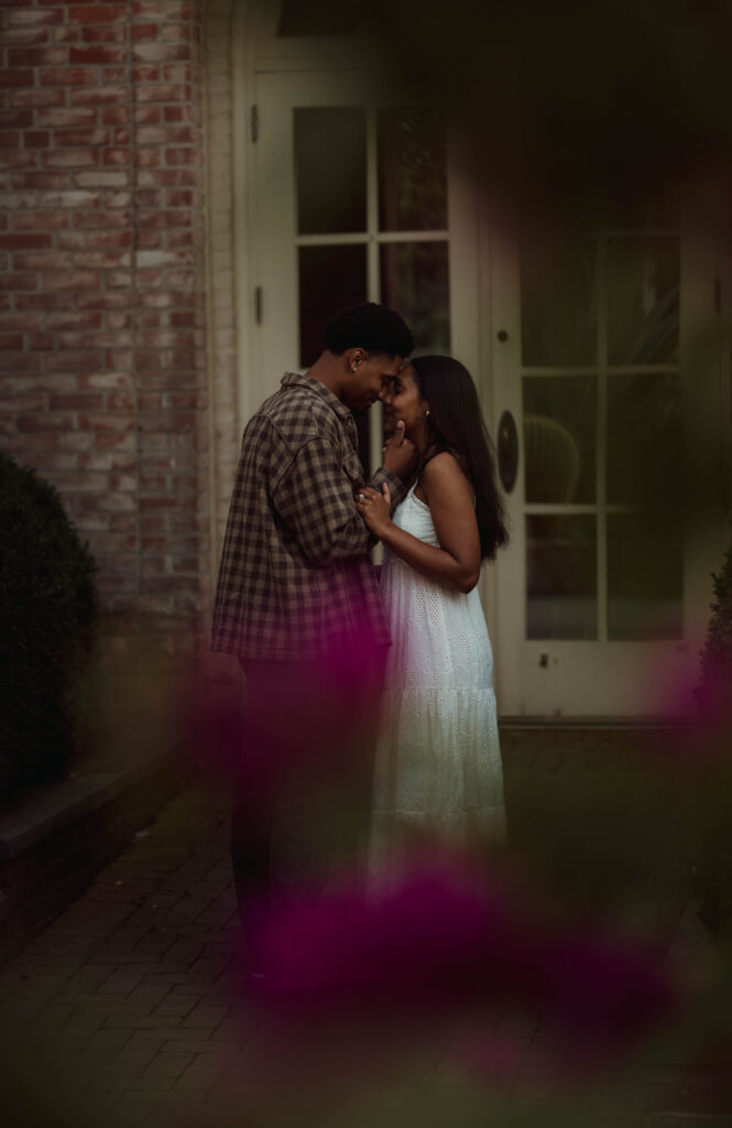 Couple embracing by a brick doorway, framed by blurred purple flowers.