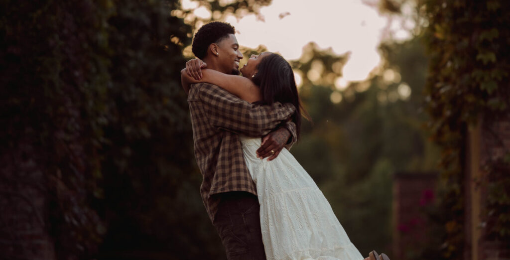 Couple embracing beside a brick building in warm evening light.