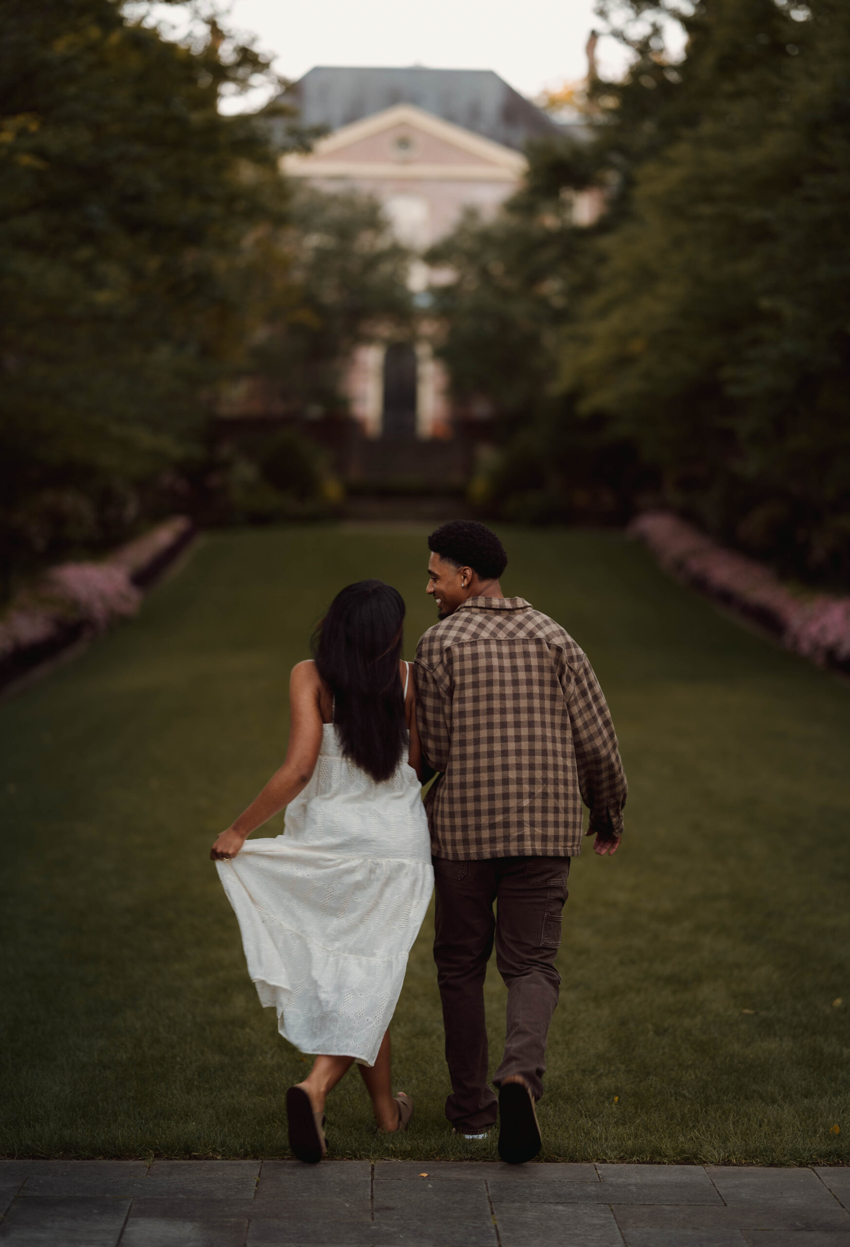 Couple holding hands and walking across a garden lawn toward a brick building.