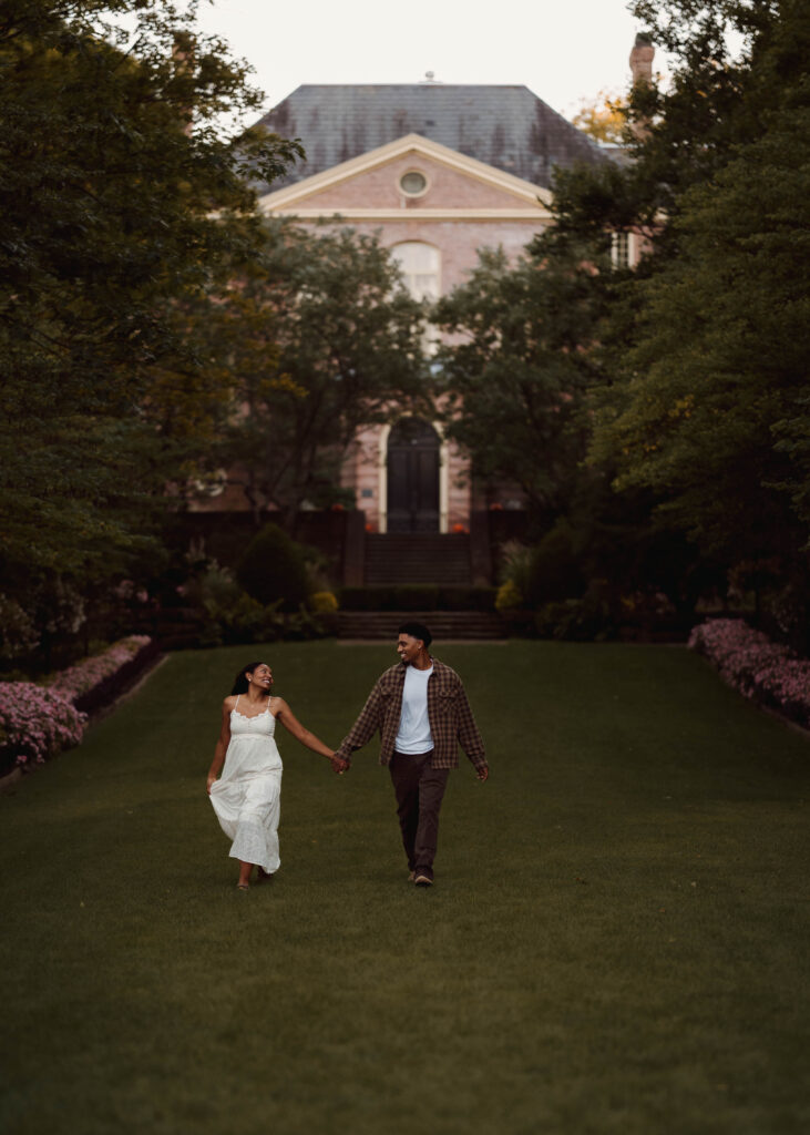 Couple holding hands while walking across a lawn in front of a brick building.